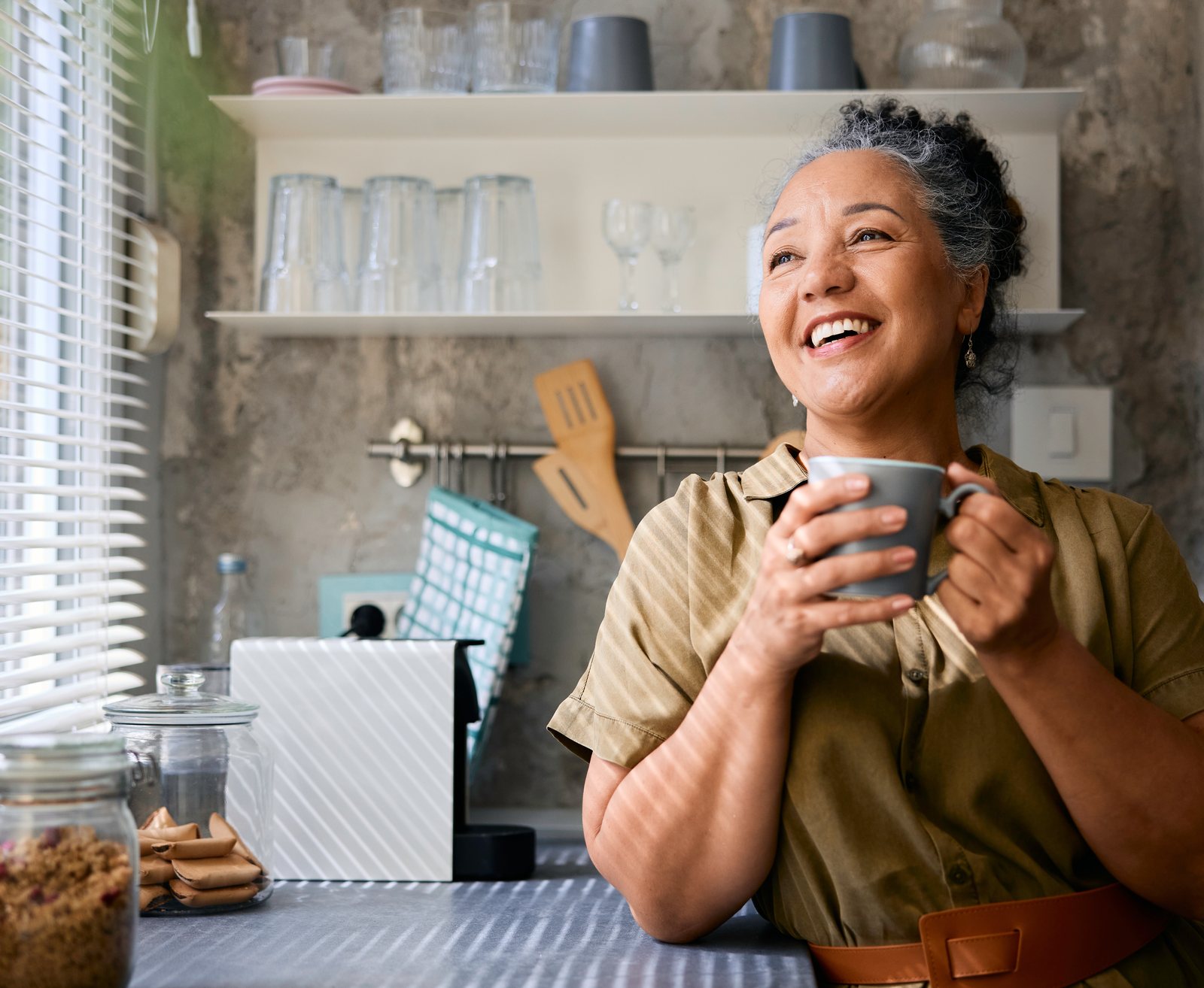A smiling older woman enjoying a cup of coffee in her sunlit kitchen.